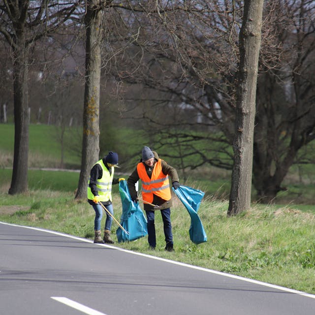 Zwei Menschen in Warnwesten und mit langen Zangen und blauen Müllsäcken gehen an einem Straßenrand und sammeln Müll.