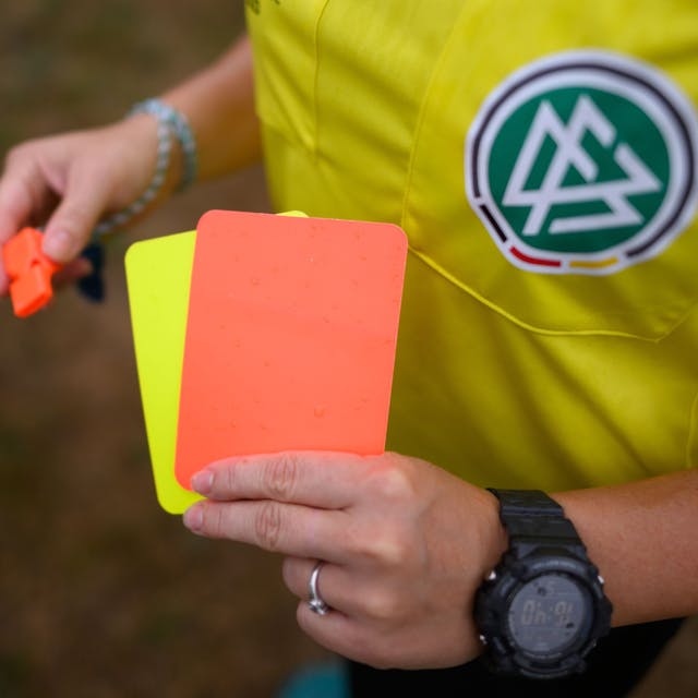 Eine Schiedsrichterin, steht mit roter und gelber Karte auf einem Rasen und hält in der anderen Hand eine Trillerpfeife.&nbsp;