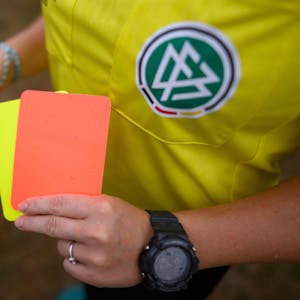 Eine Schiedsrichterin, steht mit roter und gelber Karte auf einem Rasen und hält in der anderen Hand eine Trillerpfeife.&nbsp;