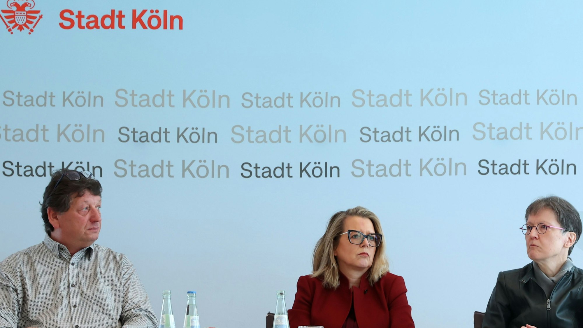 Ein Mann und Zwei Frauen sitzen an einem Tisch vor einem Plakat mit der Aufschrift Stadt Köln