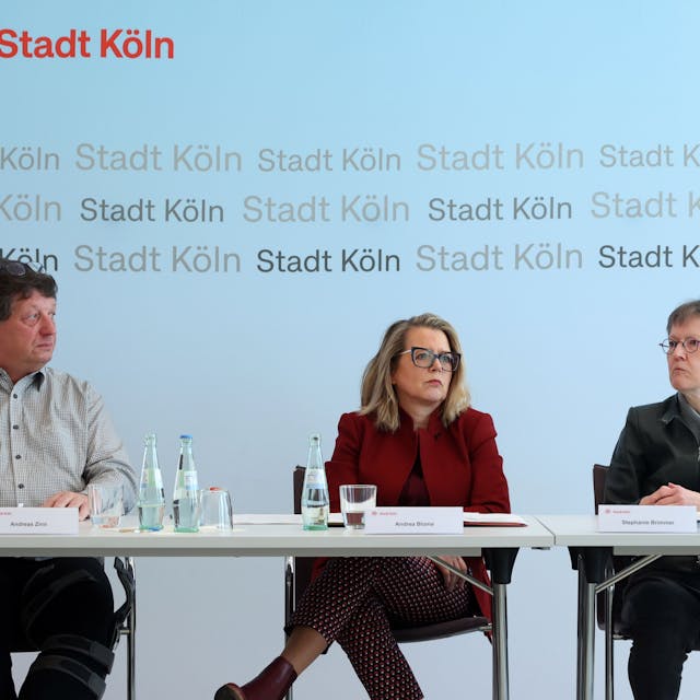 Ein Mann und Zwei Frauen sitzen an einem Tisch vor einem Plakat mit der Aufschrift Stadt Köln