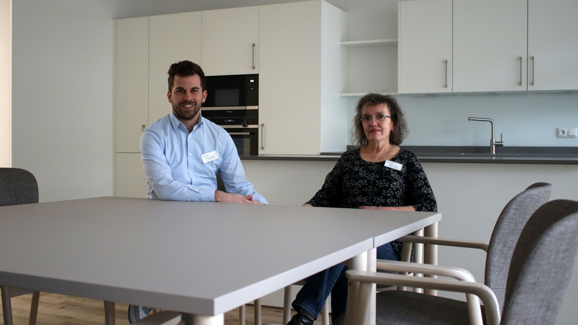 Dennis Oepen und Petra Swindt sitzen an einem Tisch der Tagespflege. Im Hintergrund befindet sich die Gemeinschaftsküche.