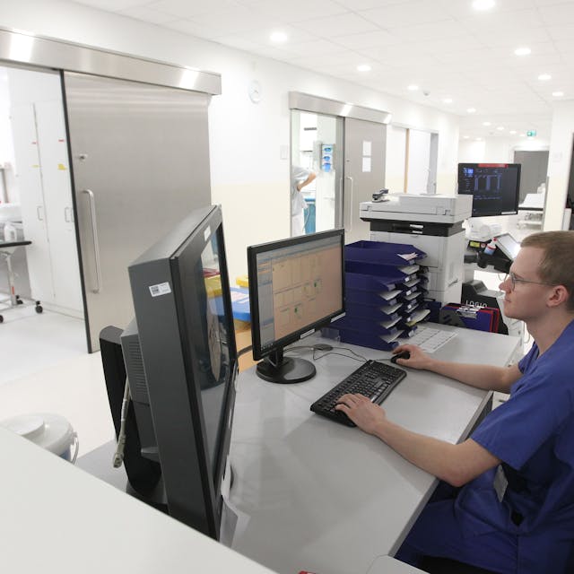Ein junger Mann in blauer Kleidung eines Pflegers sitzt an einem Schreibtisch mit zwei Bildschirmen. Im Hintergrund sind Räume mit medizinischem Gerät zu sehen.