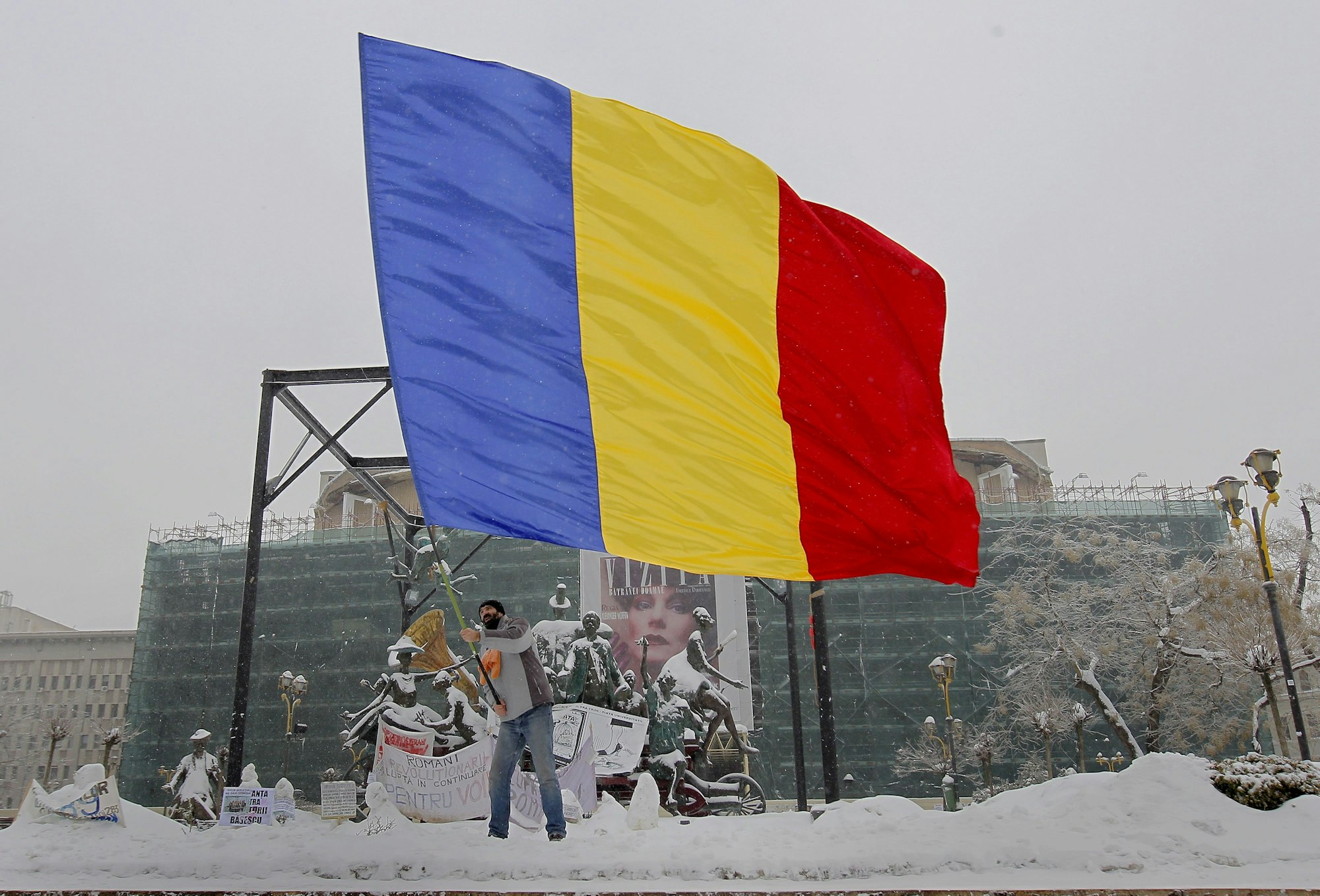 Ein Mann schwenkt eine riesige rumänische Flagge. Hinter ihm im Schnee befinden sich beschriftete Plakate.