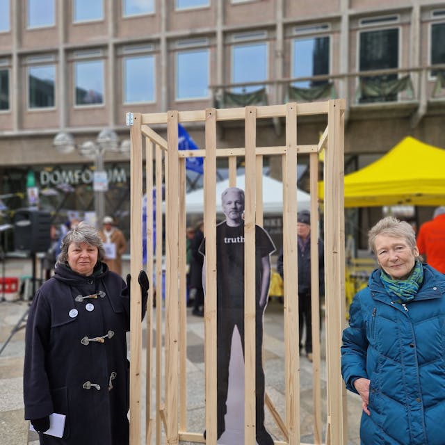 Zwei Frauen stehen zwischen einem eingegitterten Plakat von Julian Assange.