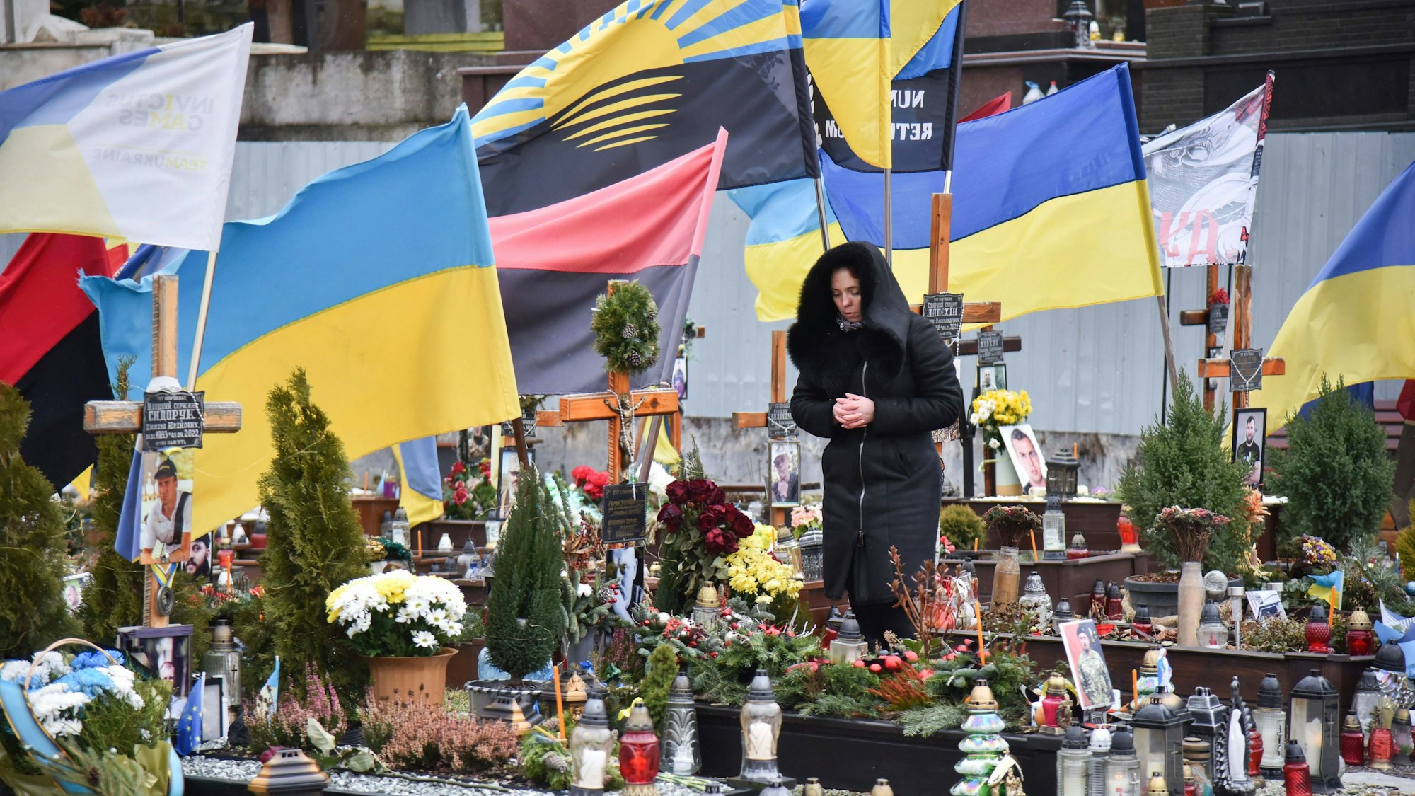 Eine Frau steht in der Nähe der russischen Stadt Lwiw auf einem Friedhof. Sie trägt schwarze Kleidung und hat die Hände gefaltet. Hinter ihr wehen ukrainische Flaggen.