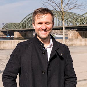 Nico Rathman steht vor dem Rheinboulevard in Köln-Deutz, im Hintergrund die Domspitzen