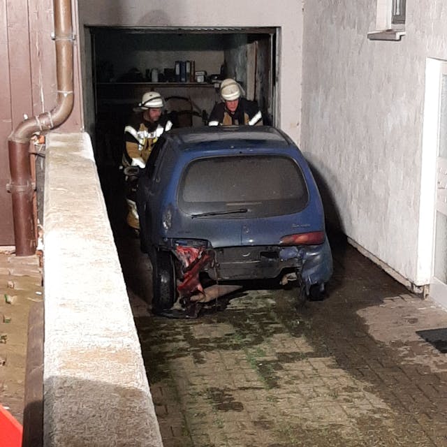 Nach einem Brand in einer Garage in Kall stehen zwei Feuerwehrleute an einem blauen Kleinwagen.