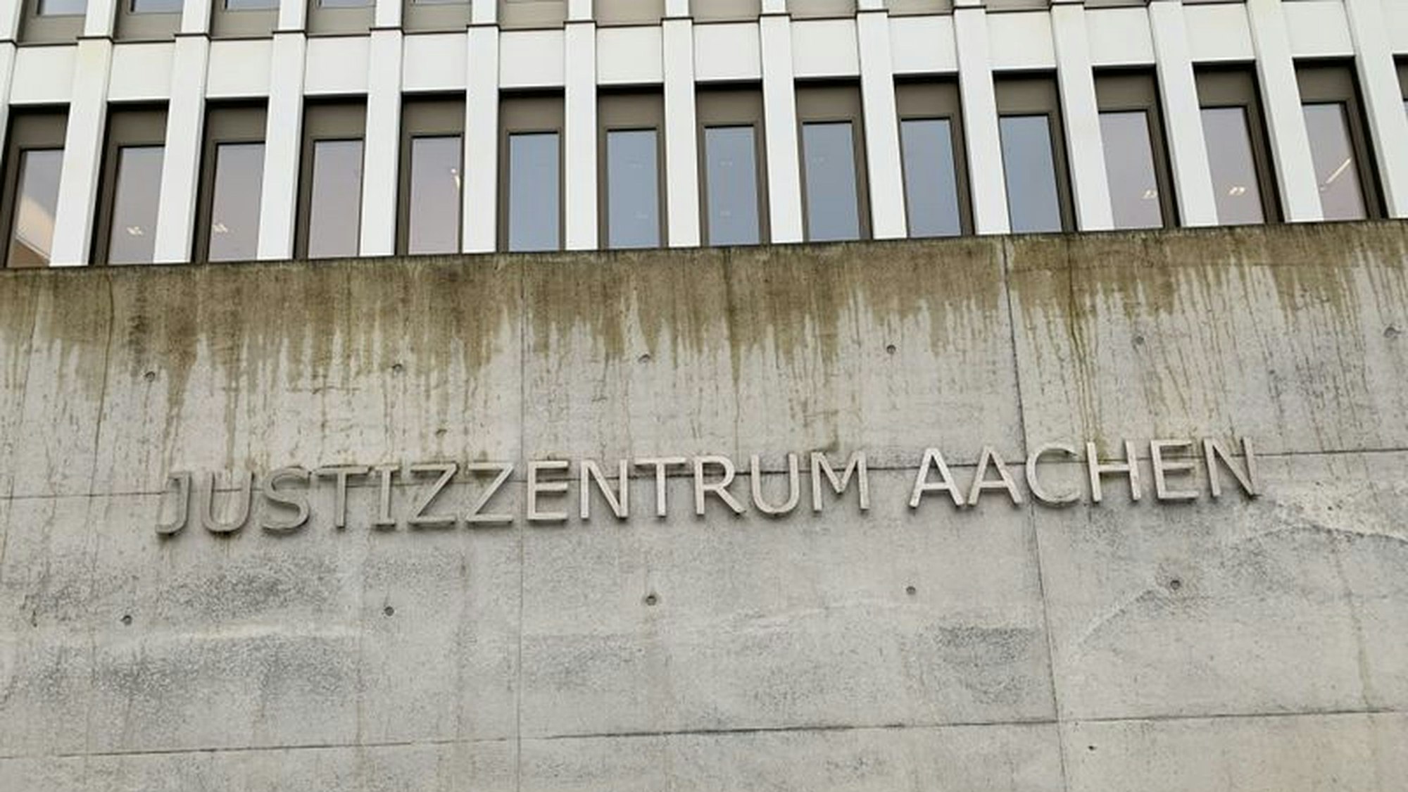 An einer Betonfassade ist der Schriftzug „Justizzentrum Aachen“ angebracht. Darüber sieht man eine Fensterreihe.