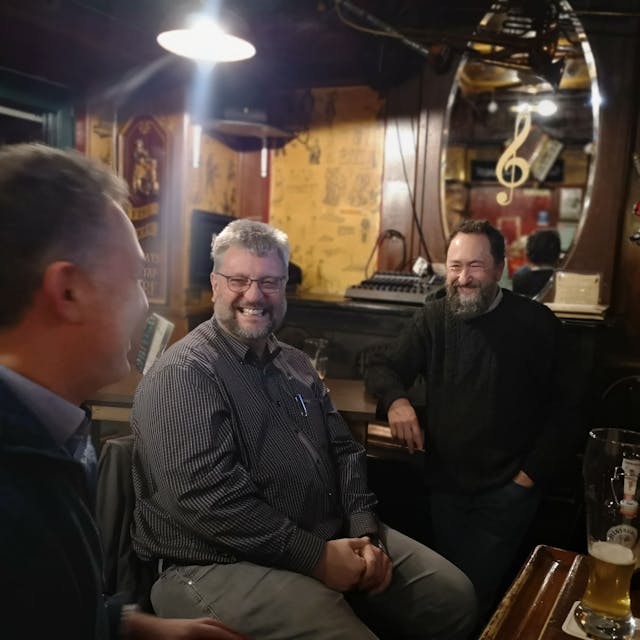 Drei Männer sitzen in einem Pub, vor ihnen stehen Biergläser auf dem Tisch, sie lachen.