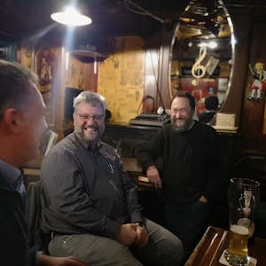 Drei Männer sitzen in einem Pub, vor ihnen stehen Biergläser auf dem Tisch, sie lachen.
