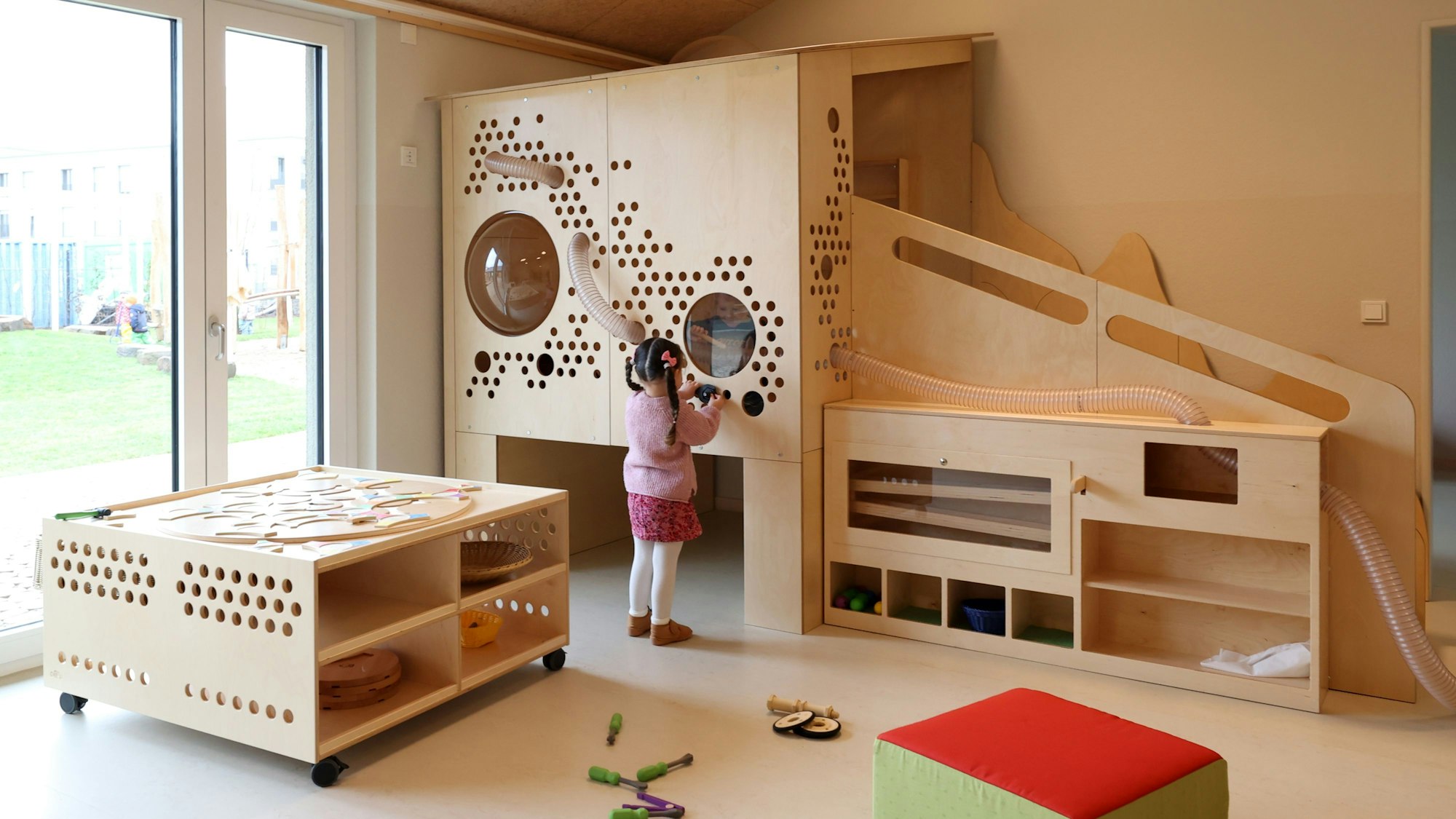 Blick in einen Gruppenraum einer Kita. Ein Kind spielt an einer Holzwand.