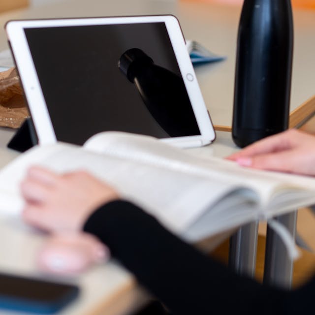 Das Bild zeigt eine Schülerin, die in einem Buch liest. Hinter dem Buch steht ein Tablet.