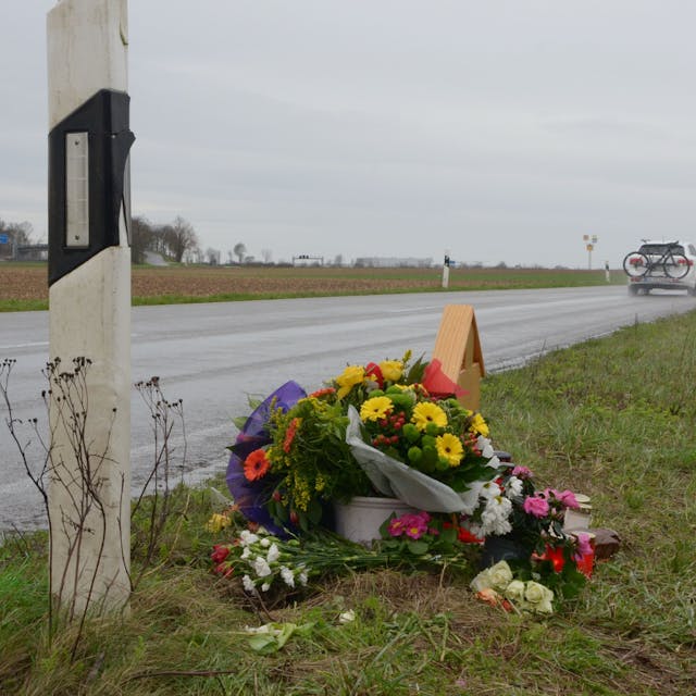 Das Bild zeigt die L163. Im Hintergrund fahren Autos, im Vordergrund steht ein Holzkreuz, das fast vollständig von Blumen verdeckt ist.