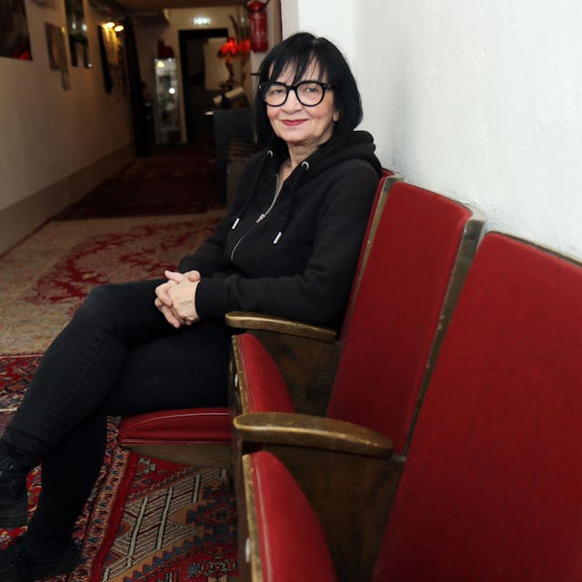 Frau mit schwarzen Haaren, schwarzer Brille und schwarzer Kleidung sitzt auf einem roten Sesel in einem Flur und schaut in die Kamera