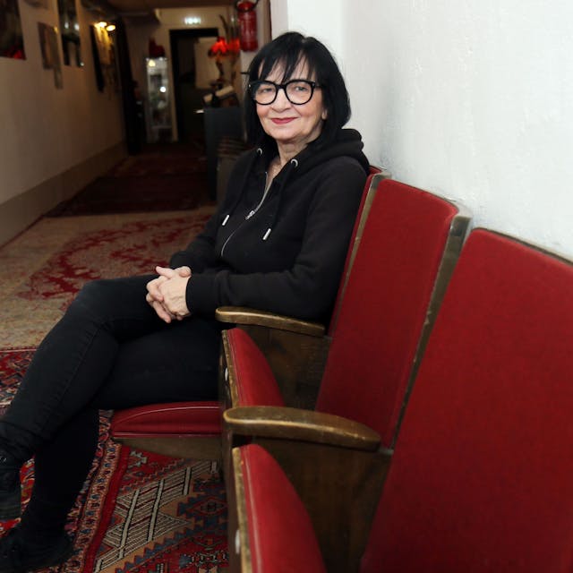 Frau mit schwarzen Haaren, schwarzer Brille und schwarzer Kleidung sitzt auf einem roten Sesel in einem Flur und schaut in die Kamera