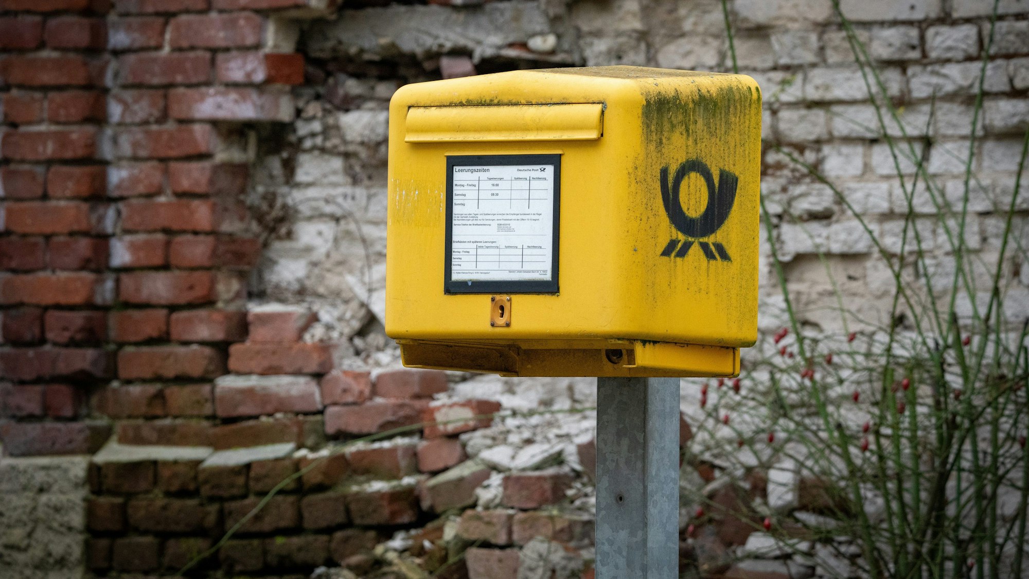 Ein gelber Briefkasten steht am Straßenrand vor einer kaputten Wand aus hellen Ziegelsteinen.