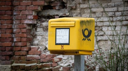 Ein gelber Briefkasten steht am Straßenrand vor einer kaputten Wand aus hellen Ziegelsteinen.