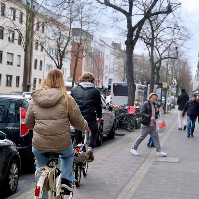Radfahrer fahren auf dem Radstreifen, Fußgänger sind auf dem direkt daneben liegenden Gehweg unterwegs.