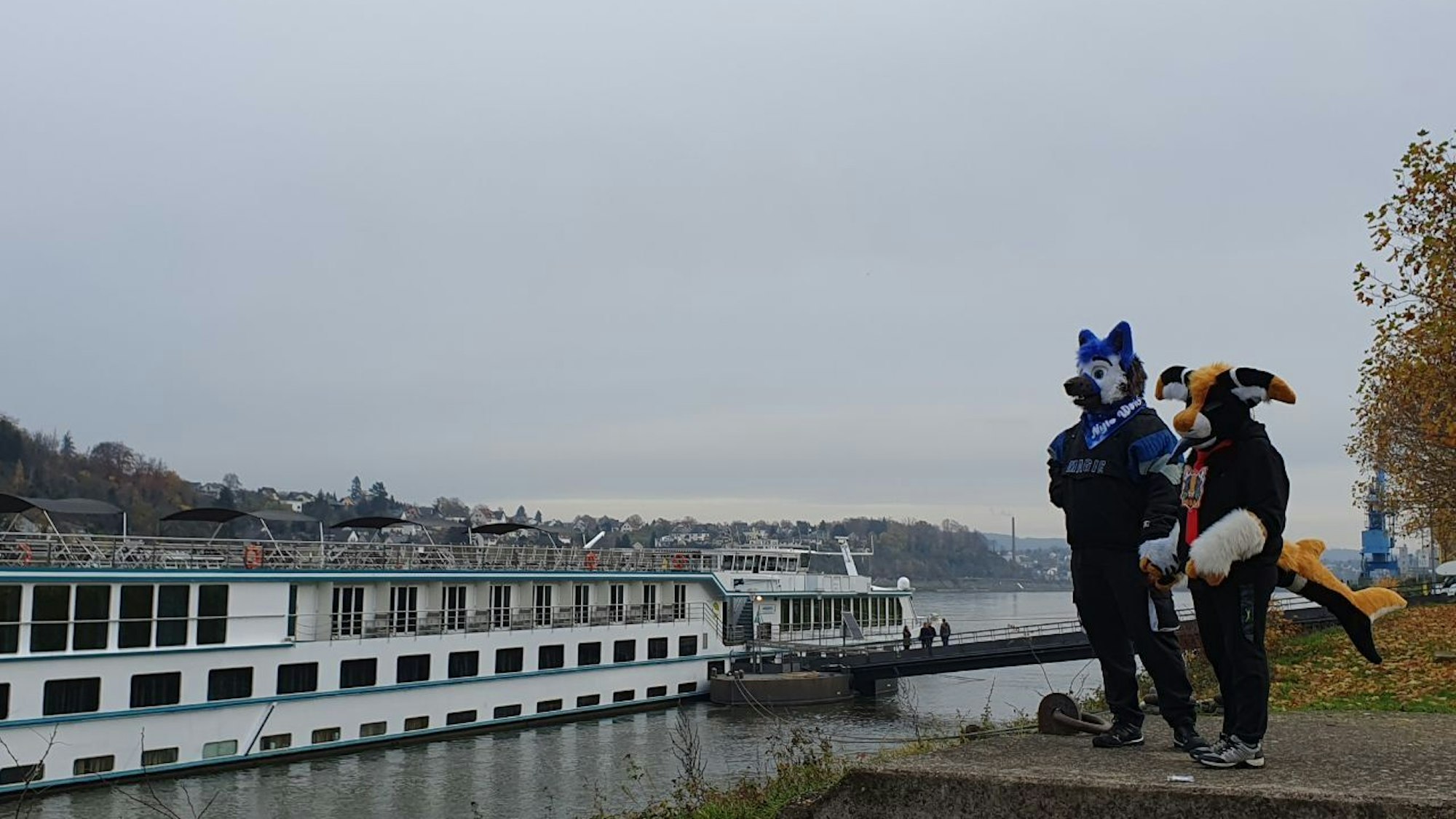Ein plüschiger Manokin und ein plüschiger Wolf stehen am Ufer eines Flusses und schauen auf die vorbeiziehenden Schiffe.