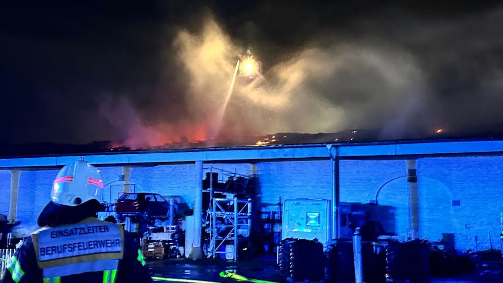 Ein Einsatzleiter der Feuerwehr steht vor einem brennenden Gebäude und beobachtet die Arbeit seiner Kollegen.