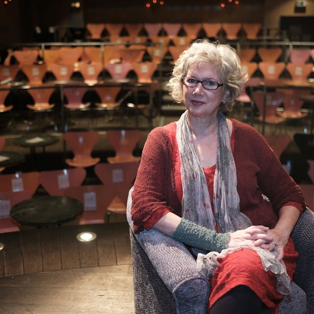 Eine Frau mittleren Alters sitzt auf der Bühne in einem kleinen Theater vor Stuhlreihen.