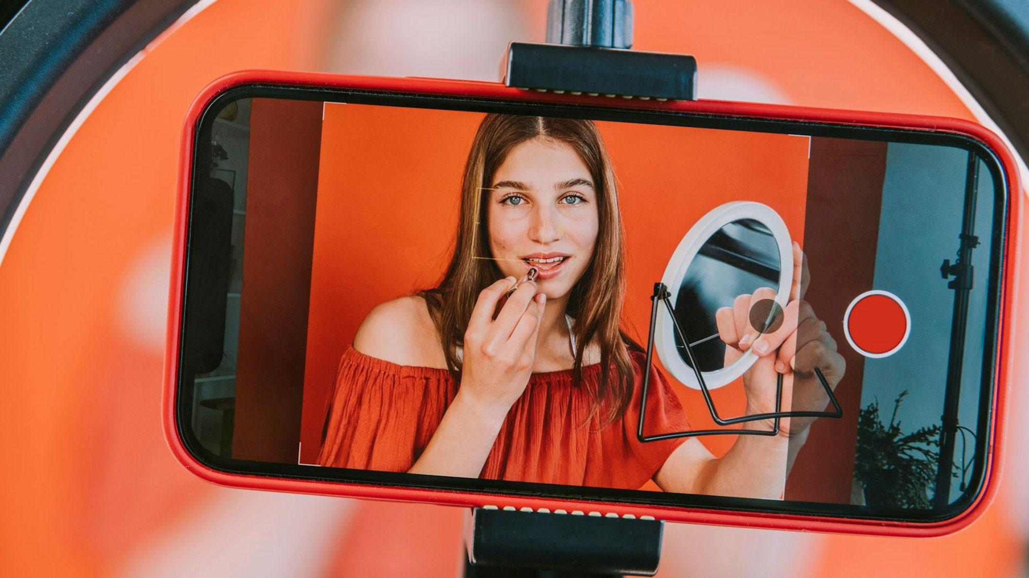 Auf einem Handy-Display sieht man das Bild einer Influencerin, die einen Spiegel in der Hand hält und sich die Lippen schminkt.