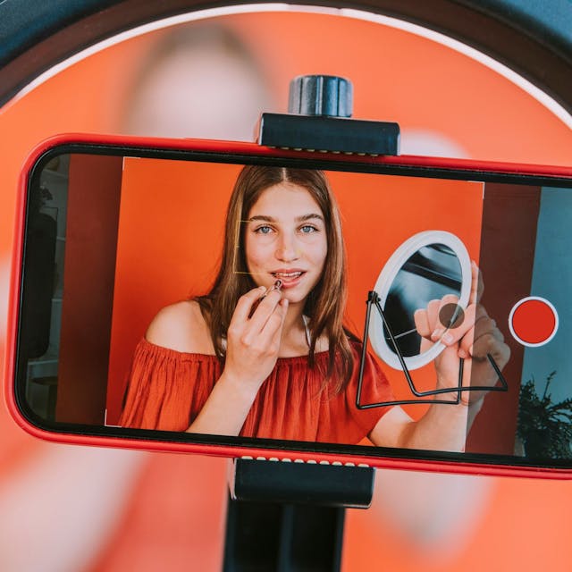 Auf einem Handy-Display sieht man das Bild einer Influencerin, die einen Spiegel in der Hand hält und sich die Lippen schminkt.