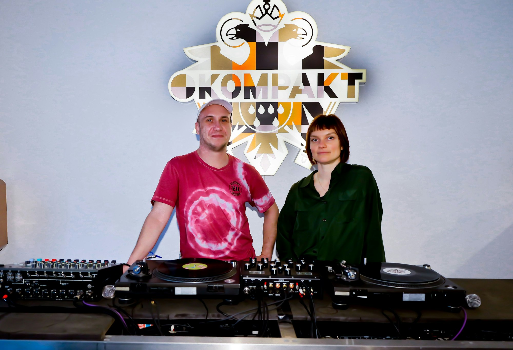 Ein Mann und eine Frau hinter einem DJ-Pult, hinter ihnen hängt das Logo des Labels Kompakt.