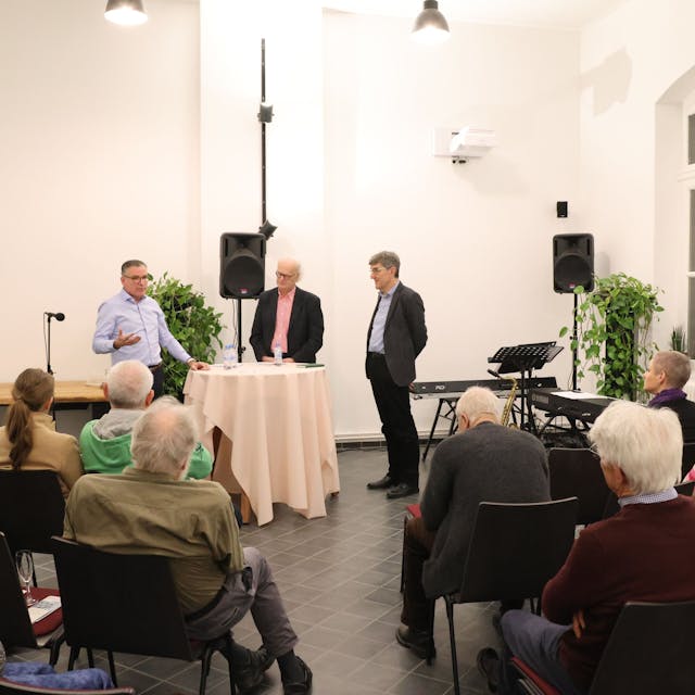 Drei Männer stehen bei einem Vortrag vor Publikum an einem Stehtisch.