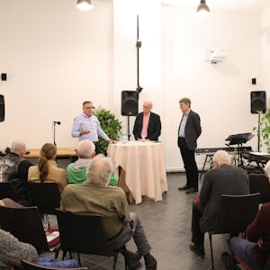 Drei Männer stehen bei einem Vortrag vor Publikum an einem Stehtisch.