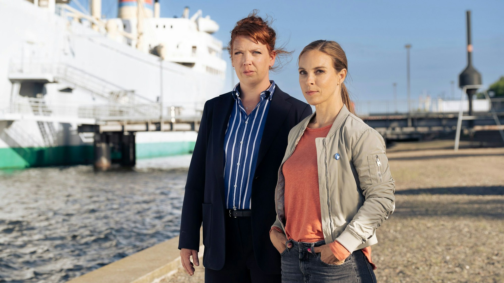 Das Bild zeigt die beiden Kommissarinnen an einem Hafen. Sie schauen ernst in die Kamera. Im Hintergrund das Wasser, ein Steg und daran ein großes Schiff.