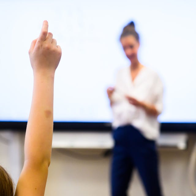 Ein Schüler meldet sich per Handzeichen, während eine Lehrerin vor einer digitalen Schultafel im Klassenraum steht.
