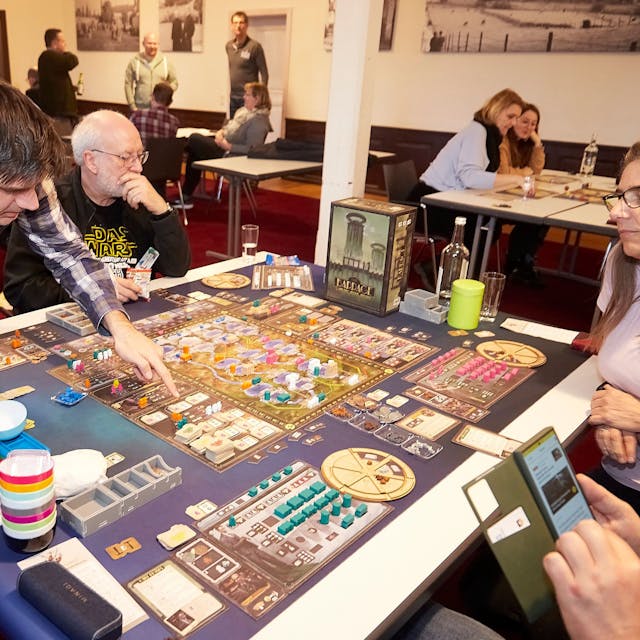 Vier Brettspieler sitzen um ein komplex aussehendes Spiel an einem Tisch. Einer von ihnen erklärt.
