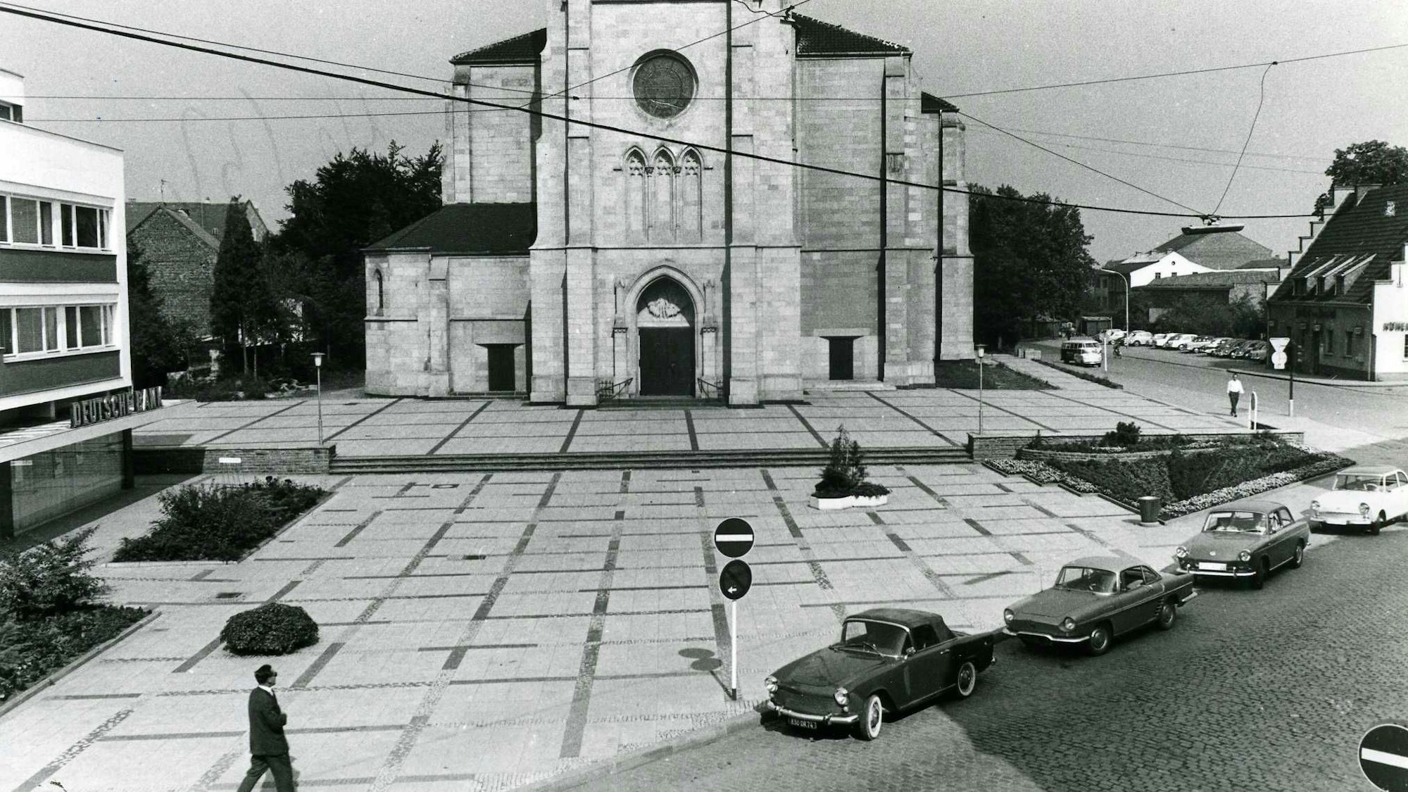 Das Bild zeigt eine historische Aufnahme des Herz-Jesu-Vorplatz. Im Vordergrund sind alte Autos zu erkennen.