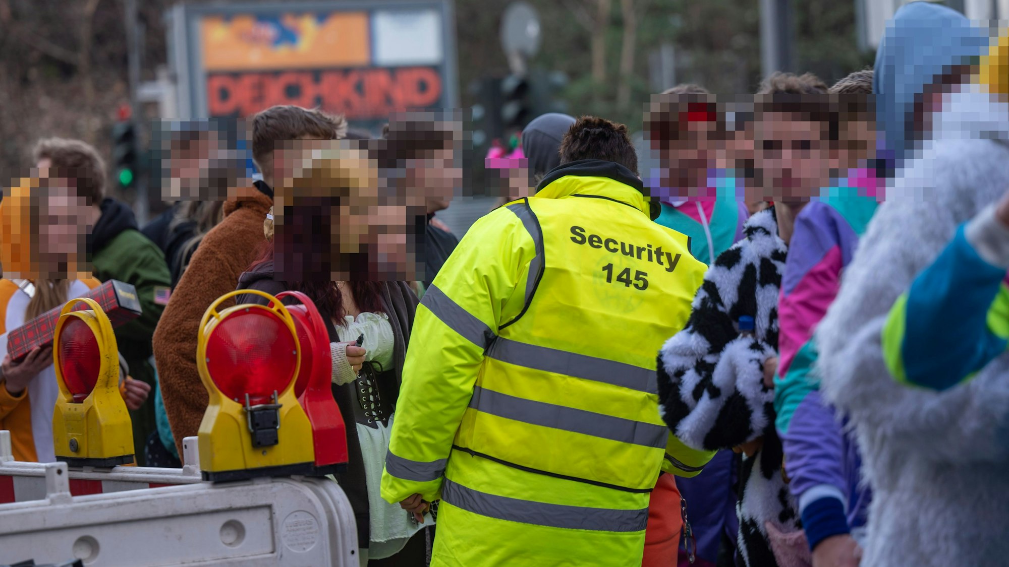 Zu sehen sind eine Gruppe von Menschen. Ein Security-Mitarbeiter in einer gelben Jacke ist in der Rückansicht zu sehen.