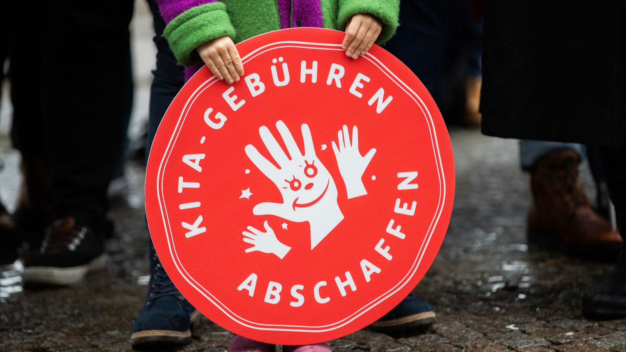 Ein Kind hält ein rotes, rundes Plakat mit der Aufschrift "Kita-Gebühren abschaffen" vor sich.