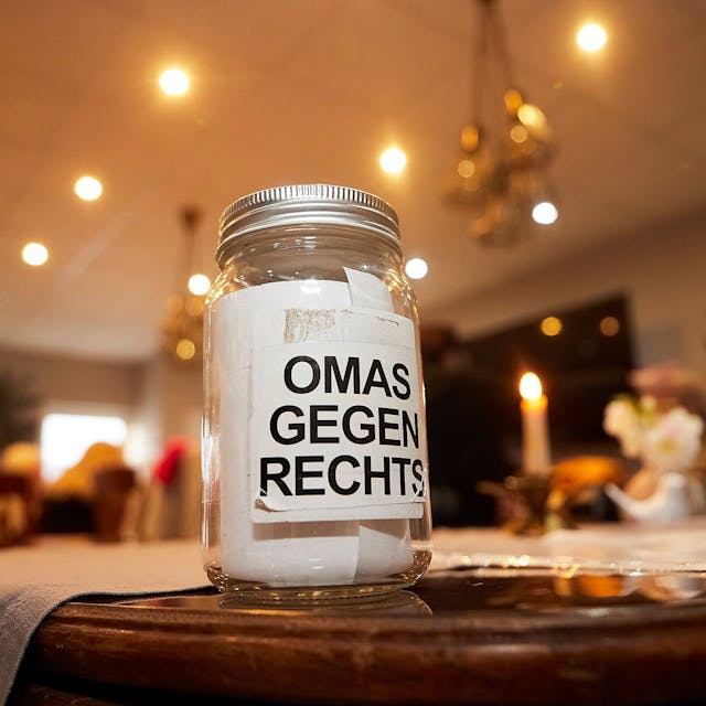 Ein Glas mit der Aufschrift Omas gegen Rechts steht auf einem Tisch. Im Hintergrund sind eine brennende Kerze und Menschen an weiteren Tischen zu erahnen.