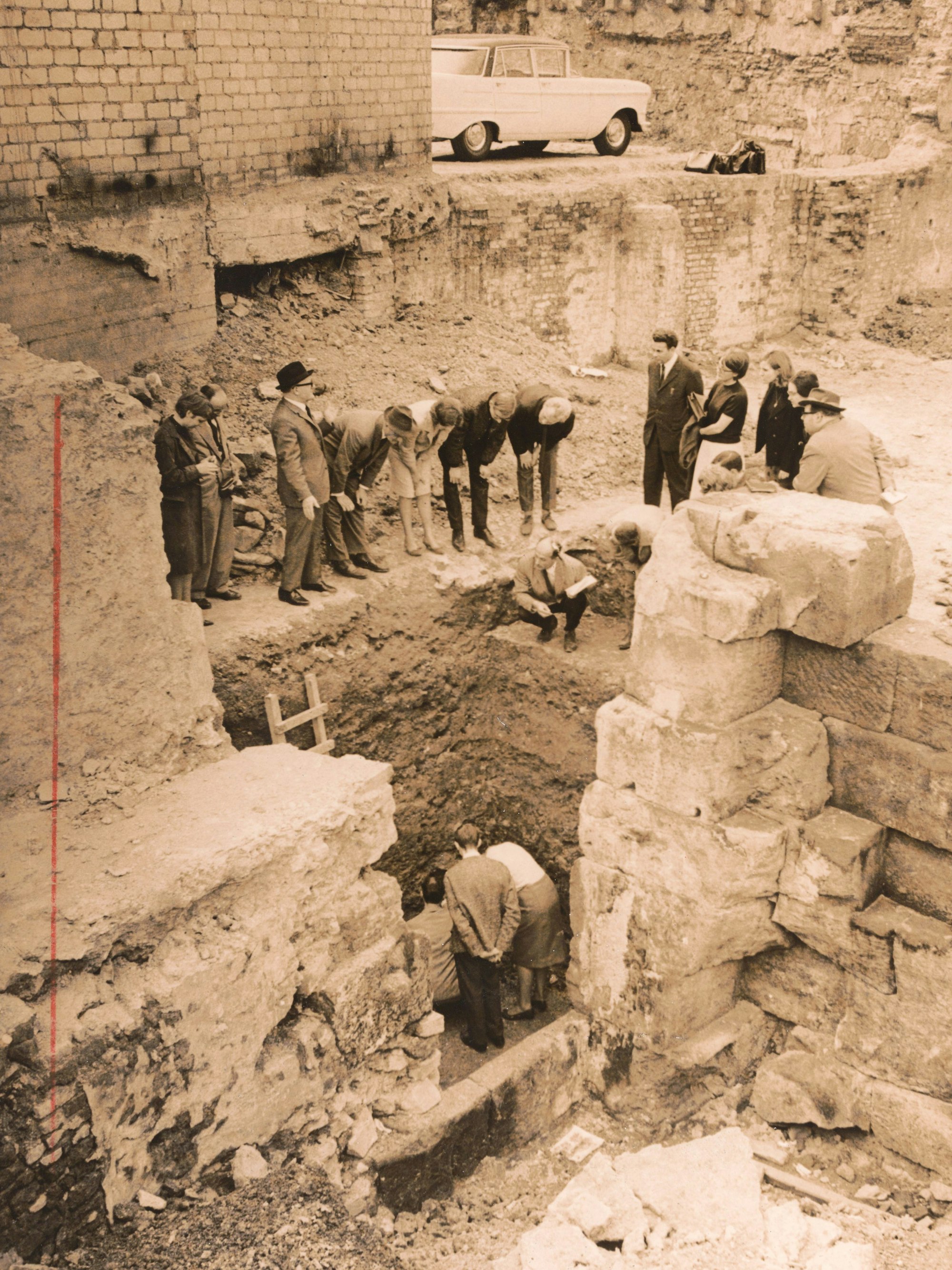 Bei Arbeiten für den Dombunker wurde das Dionysosmosaik entdeckt. Das Foto wurd bei der Ausgrabung im August 1970 aufgenommen. Schick gekleidete Personen stehen vor einer Grube zwischen alten Mauern, zwischen denen das Mosaik entdeckt wurde.