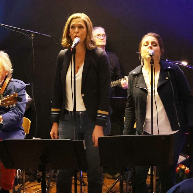 Die drei Sängerinnen stehen nebeneinander im Vordergrund, die Instrumentalisten sitzen dahinter.