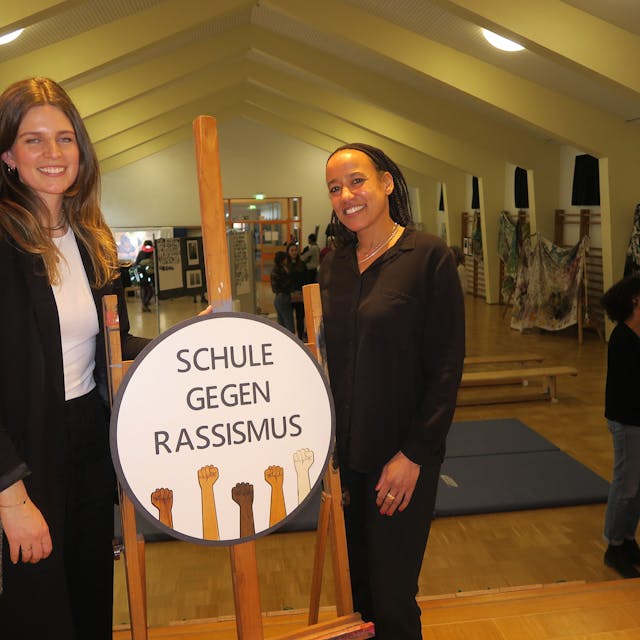 In einer Turnhalle stehen zwei junge Frauen bei einem Aufsteller, auf dem Schule gegen Rassismus steht