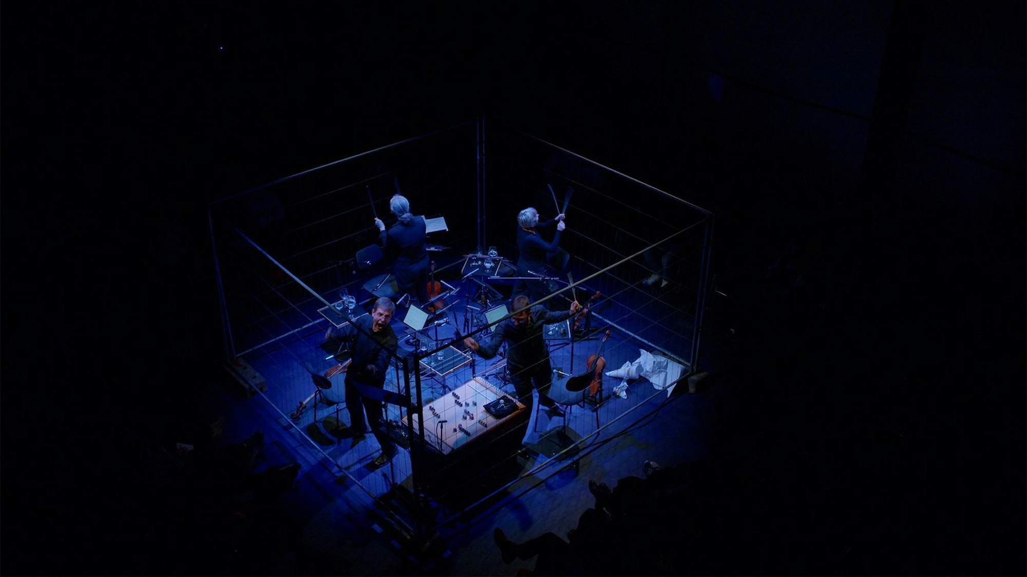 Zu sehen sind vier Musiker mit ihren Instrumenten in der Mitte des bildes, eingepfercht mit Plexiglasscheiben und bläulichz beleuchtet. Der Rest des Bildes ist schwarz.