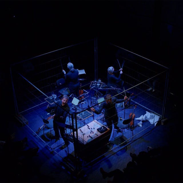 Zu sehen sind vier Musiker mit ihren Instrumenten in der Mitte des bildes, eingepfercht mit Plexiglasscheiben und bläulichz beleuchtet. Der Rest des Bildes ist schwarz.&nbsp;