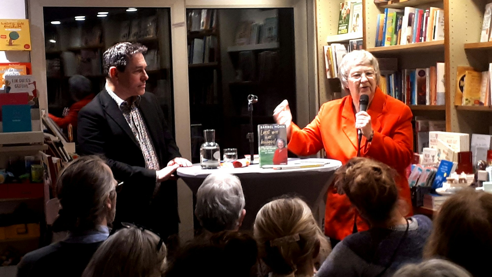 Ein Mann und eine Frau im orangen Blazer stehen in einer Buchhandlung vor Publikum an einem Stehtisch.