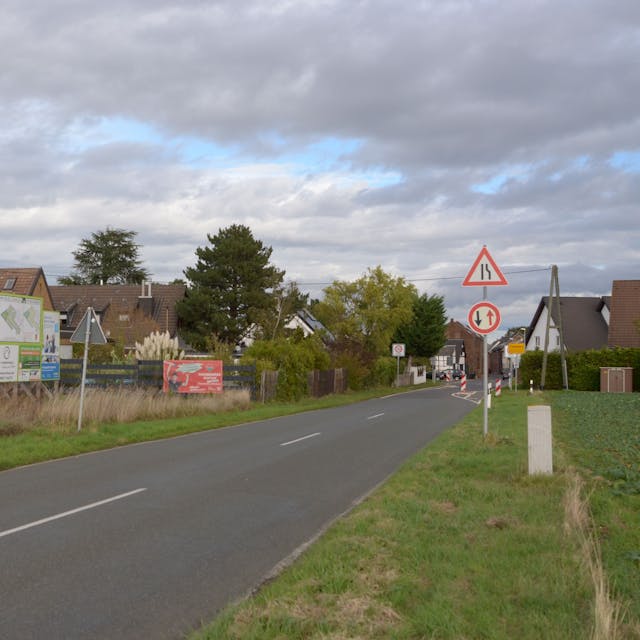 Das Bild zeigt die Nideggener Straße, die nach Frauenberger hineinführt. Am Straßenrand steht eine Werbetafel, die auf das geplante Wohngebiet aufmerksam macht.