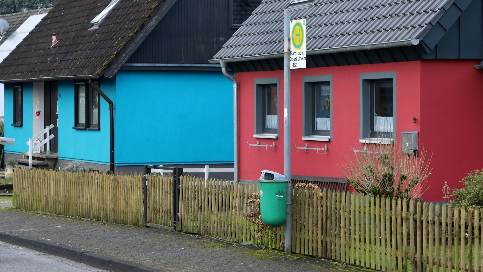 Die einsamste Bushaltestelle Bergisch Gladbachs steht in Oberkühlheim. Ein Mülleimer und ein Bushaltestellenschild stehen vor einem roten Haus, weiter hinten links im Bild ist ein türkises Haus.