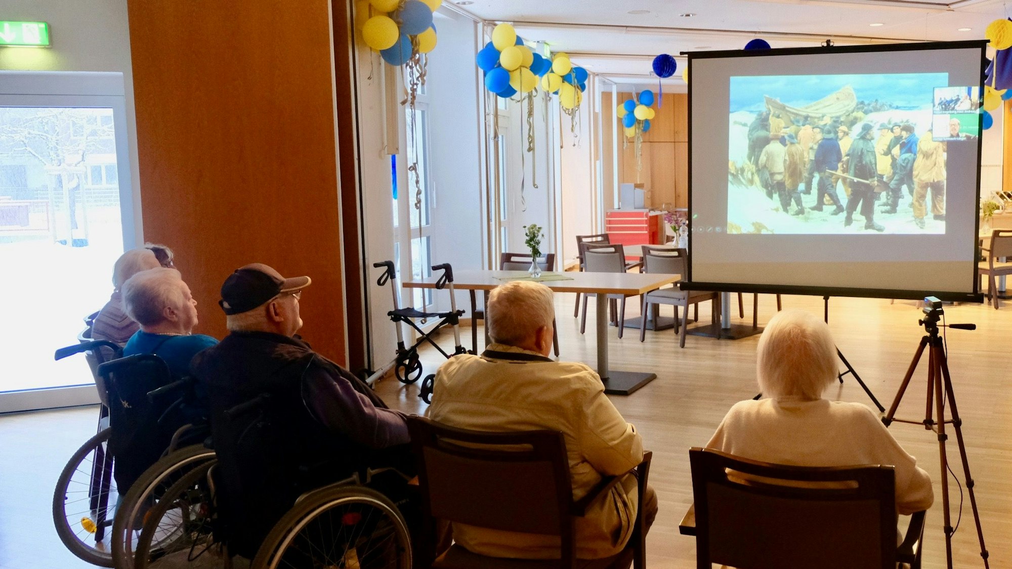 Einige ältere Menschen sitzen im Halbkreis in einem großen Saal. Sie betrachten ein Bild, das auf eine Leinwand projiziert wird.