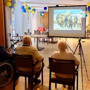 Einige ältere Menschen sitzen im Halbkreis in einem großen Saal. Sie betrachten ein Bild, das auf eine Leinwand projiziert wird.