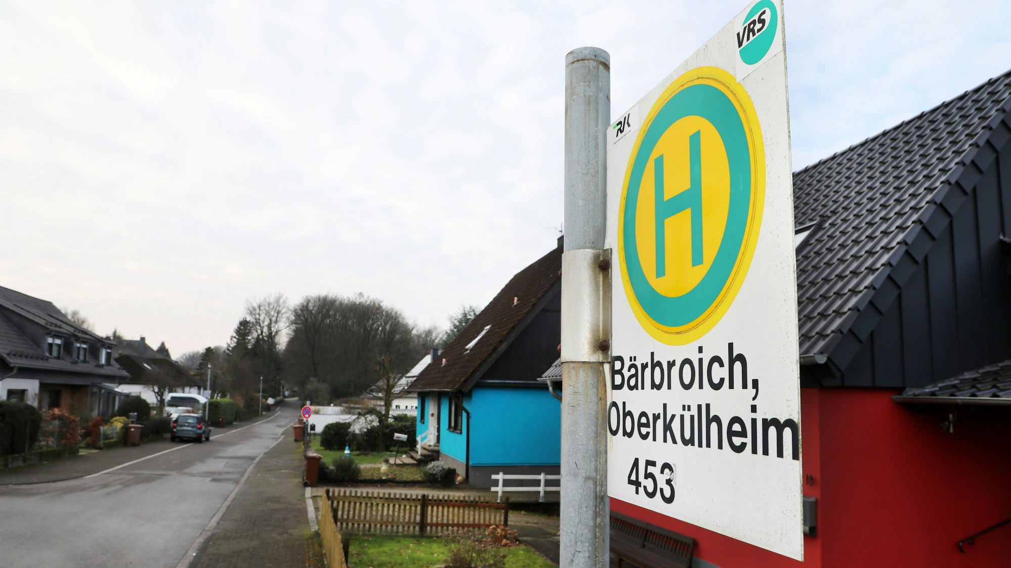 Das Haltstellenschild der Bushaltestelle „Bärbroich, Oberkühlheim“ in Bergisch Gladbach ist zu sehen, im Hintergrund eine Wohnsiedlung mit kleinen Vorgärten.