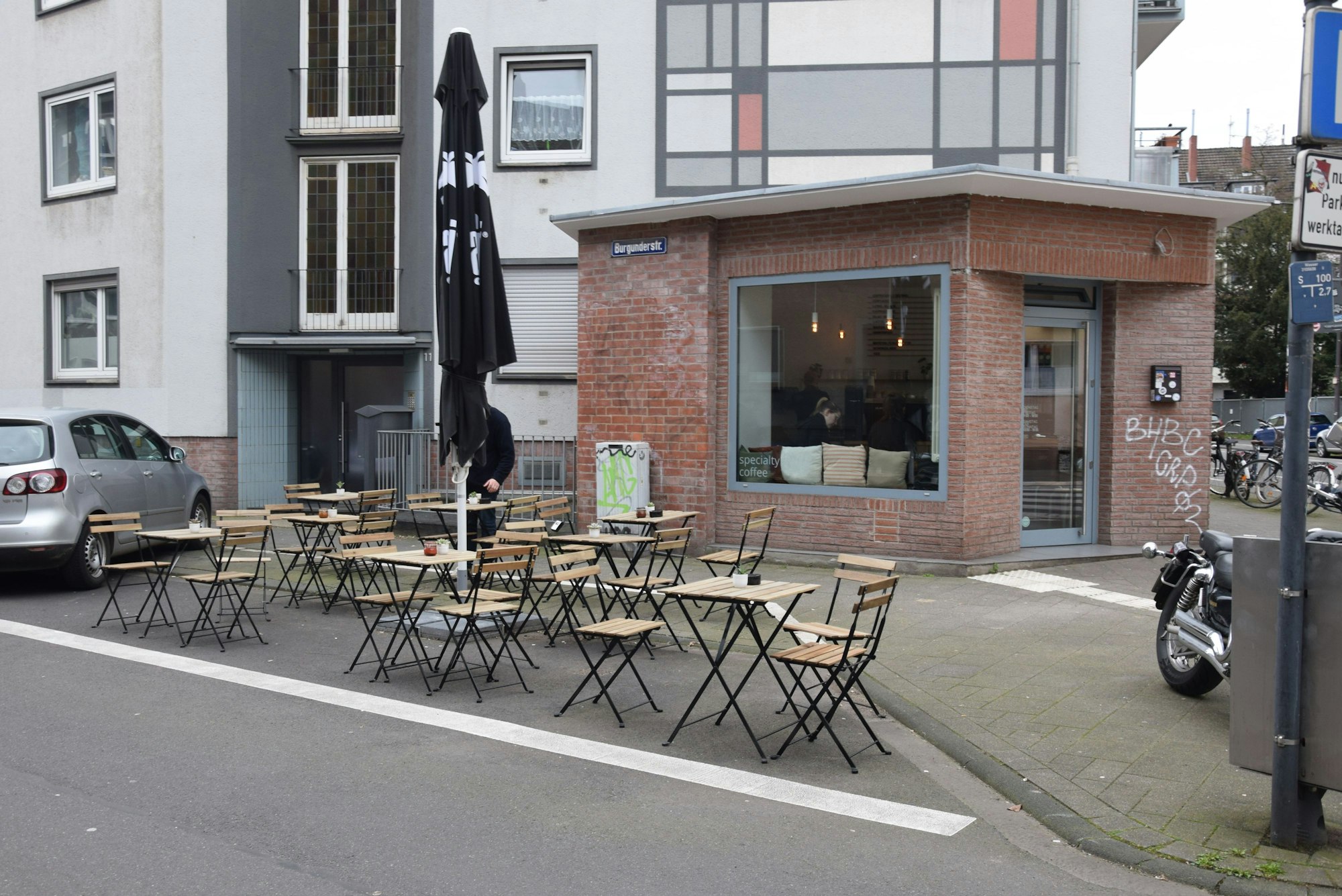 Auf einem Parkplatz stehen Tische, die zu einem dahinterliegenden Café in einem schlichten Eckhaus gehören.